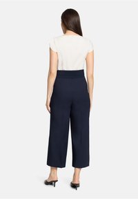 Femme aux longs cheveux bruns portant un chemisier blanc à manches courtes, un pantalon large taille haute bleu marine et des mules à talons noires, debout, vue de dos.