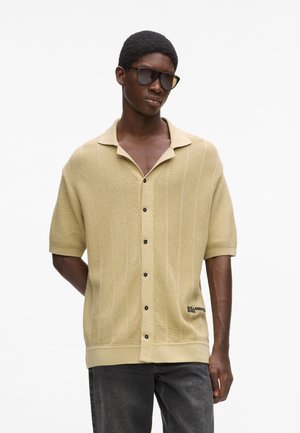 Lühikeste varrukatega, helekollane kootud kardigan tekstuurse mustriga, nööpidega kinnitatav ja musta logoga detail alumisel esiküljel.