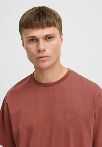 Brun bomulds t-shirt med rund halsudskæring, korte ærmer og et tonalt broderet cirkellogo på venstre bryst. Glat tekstur.