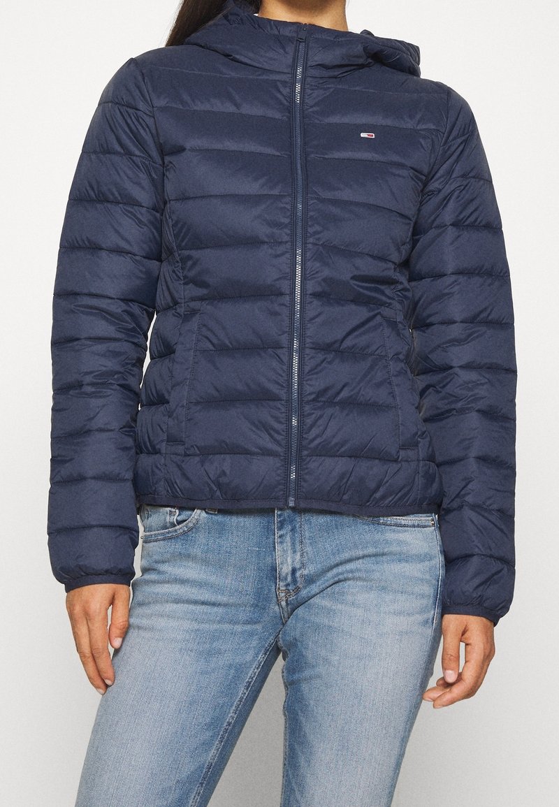 Marinblå pufferjacka med huva, försedd med horisontell quiltning, en framdragkedja och en liten logotyp på bröstet. Lämplig för kallt väder.