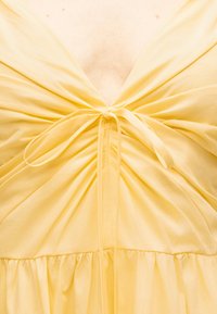 Κίτρινο φόρεμα με βαθύ V-λαιμόκοψη, ρυτιδωμένο μπροστινό μέρος και δεμένο φιόγκο στο κέντρο του στήθους.