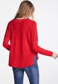 Rotes langärmliges Strickoberteil mit Rippstruktur, runden Ausschnitt und Seitenschlitzen, kombiniert mit blauen Jeans für einen lässigen Look.