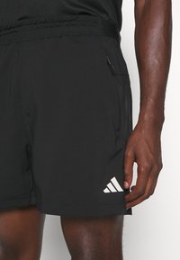 Svarta idrottsshorts med elastisk midja, ficka med dragkedja och en vit tre-randig logotyp på nedre vänstra sidan. Slät yta.