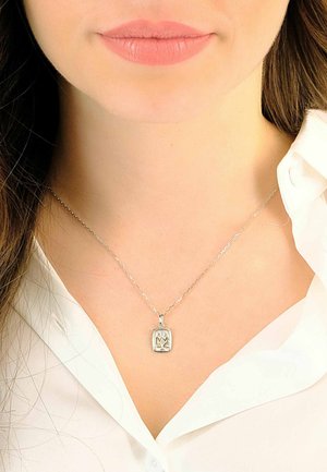 Collana d'argento con un pendente rettangolare decorato con un motivo floreale, appoggiata su una camicia bianca con colletto.