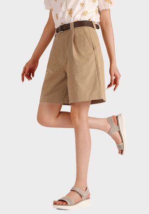 Donna che indossa pantaloncini beige fino al ginocchio con cintura marrone, camicetta floreale e sandali aperti grigio chiaro, in piedi su una gamba piegata.