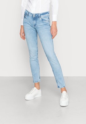 Person iført lyseblå slim-fit jeans, hvid langærmet skjorte og hvide platformsneakers, der står på et ensartet gulv.