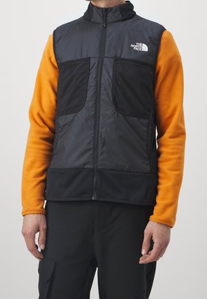 Mężczyzna w czarnej kamizelce na zamek, z pomarańczowymi długimi rękawami i czarnych spodniach, kamizelka ma logo The North Face na klatce piersiowej.