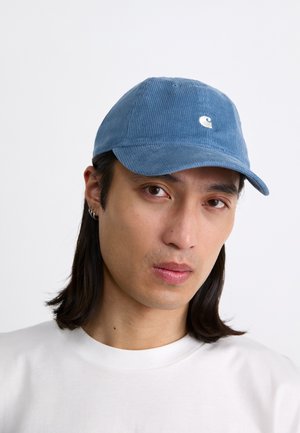 Boné de veludo azul com aba curva e logótipo bordado na frente, usado por uma pessoa com uma camisa branca. Tecido texturizado e design casual.