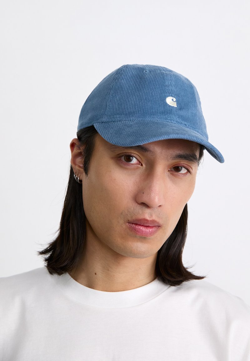 Boné de veludo azul com aba curva e logótipo bordado na frente, usado por uma pessoa com uma camisa branca. Tecido texturizado e design casual.