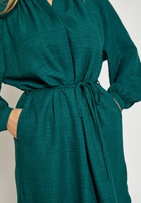 Obleka za zavijanje v zeleno-modri barvi iz teksturirane tkanine, z dolgimi rokavi, pasom in stranskimi žepi.