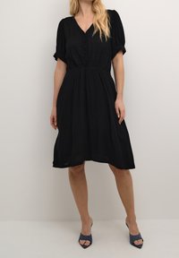 Schwarzes Kleid mit V-Ausschnitt, kurzen Puffärmeln und Knopfdetails an der Vorderseite. Luftiger Stoff und ausgestellte Silhouette. Getragen mit blauen offenen Pumps.