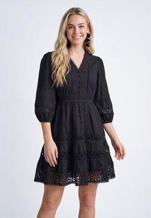 Junge Frau mit langen blonden Haaren trägt ein schwarzes Kleid mit Spitze, dreiviertel Ärmeln und großen schwarzen Blumenohrringen, lächelnd.