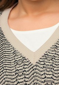 Krämsfärgad stickad tröja med V-ringning och ett texturerat mönster i svart och kräm. Urringningen är accentuerad med en ljusbeige kant.