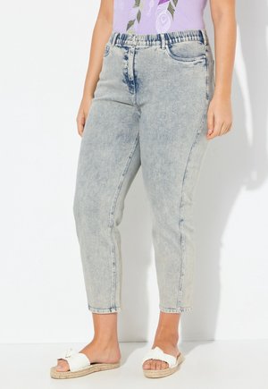 Jean court délavé clair avec taille élastique porté par une personne debout, associé à des sandales blanches à enfiler et à un haut lilas avec motif feuilles.