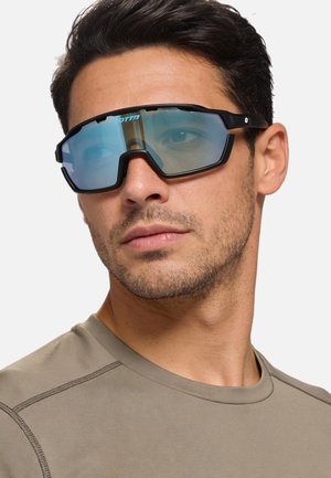 Uomo che indossa occhiali sportivi oversize con montatura nera e lenti azzurre e una maglietta beige a girocollo, che guarda leggermente di lato.