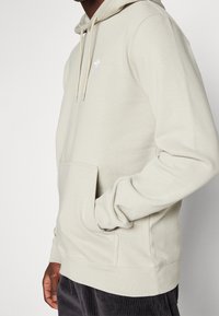 Muž v béžové mikině Adidas s přední kapsou a logem, ruka zasunutá v kapse, v kombinaci s tmavými kordovou kalhotami.