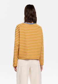 Langarm, mosterdgeel gestreept shirt met roze strepen, met een blauwe ronde hals en een relaxte pasvorm. Gedragen met lichtcrème gekleurde broeken.