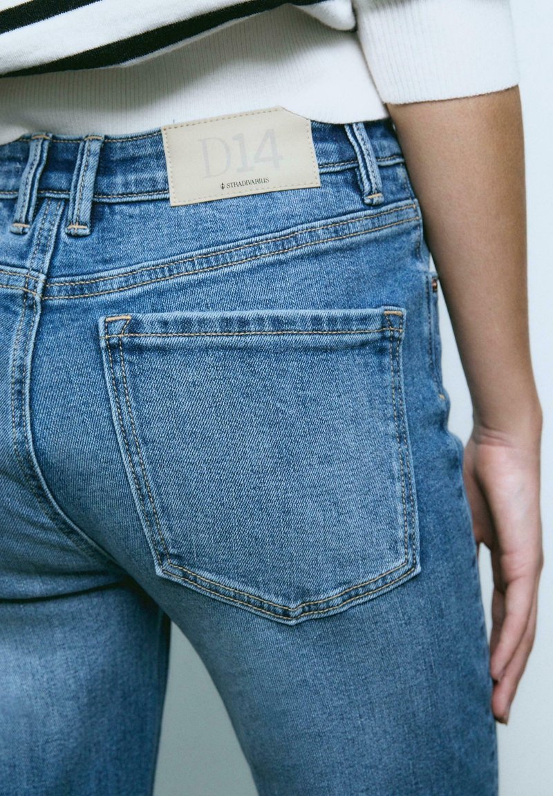 Gros plan sur une personne portant un jean en denim bleu avec une poche arrière et une étiquette beige marquée "D14" et "STRADIVARIUS" à la taille.