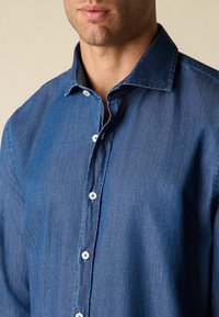 Mężczyzna w średnio niebieskiej koszuli dżinsowej z guzikami i kołnierzykiem, pokazany od ramion do ust na jednolitym tle.