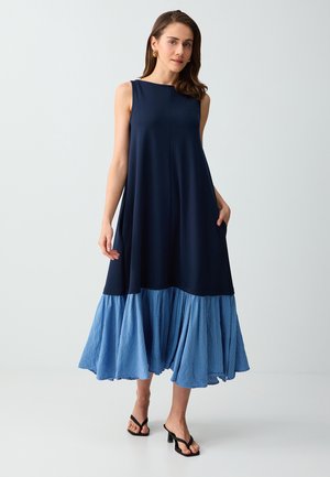 Femme portant une robe midi sans manches bleu marine avec un ourlet plissé à rayures bleues et blanches, des sandales noires à talons, debout devant un fond clair.