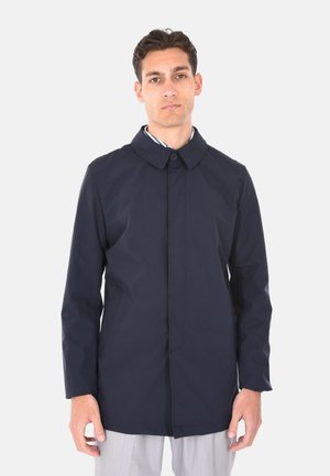 Giovane uomo che indossa un cappotto monopetto blu navy scuro con colletto sopra pantaloni grigio chiaro, in piedi contro uno sfondo chiaro uniforme.
