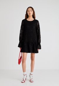 Μαύρο δαντελένιο φόρεμα με μακριά, διάφανα μανίκια και βολάν στο τελείωμα. Το μοντέλο κρατά μια κόκκινη τσάντα και φορά λευκές κάλτσες με σανδάλια κόκκινου τακουνιού.