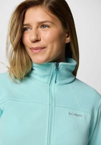 Giacca in fleece azzurro chiaro con colletto alto, chiusura con zip e logo. Texture morbida e dettaglio delle cuciture visibile sul davanti.