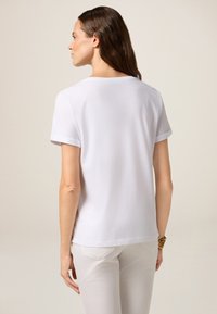 Vrouw met lang bruin haar, gekleed in een eenvoudige witte T-shirt met korte mouwen en witte broek, staand tegen een neutrale achtergrond, met de rug naar de camera.