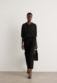 Zwarte blouse met strikdetail, zwarte op maat gemaakte broek en zwarte puntige hakken. Model draagt een kleine zwarte handtas en staat op een neutrale vloer.