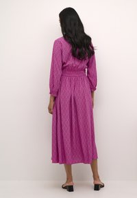 Duga, ružičasta haljina s uzorkom, dugim rukavima, skupljenim strukom i lepršavom suknjom, napravljena od lagane tkanine. Crne sandale s blok petom.