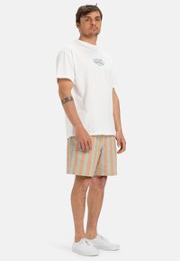 White cotton t-shirt with a light blue wave and sun graphic, paired with striped shorts in teal and peach. White sneakers complete the look.