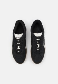 Zapatillas de ante negro con acentos blancos, que presentan un diseño de cordones, parte superior texturizada y suela de goma marrón. Se aprecia el logo de Reebok.