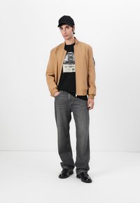 Tan bomberjakke med elastiske mansjetter, svart grafisk t-skjorte, grå rette jeans og svarte sko, med en svart caps som fullfører antrekket.