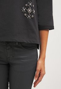 Sudadera negra con mangas 3/4, adornada con cuentas plateadas y perlas en un patrón floral. Combinada con jeans oscuros.