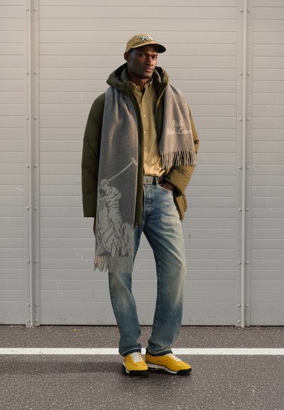 Polstret olivengrønn jakke, tan farge knappet skjorte, grå skjerf med frynser og polodesign, falmede blå jeans og gule joggesko med hvite detaljer.