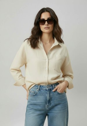 Femme aux cheveux bruns ondulés portant de grandes lunettes de soleil noires, une chemise crème boutonnée et un jean bleu taille haute, posant les mains dans les poches.