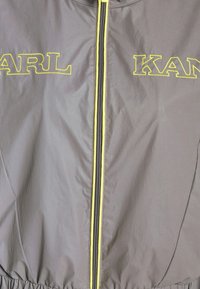 Szara lekka kurtka z żółtymi akcentami, z zamkiem z przodu i haftem "KARL" oraz "KAN" na klatce piersiowej w wyrazistym stylu.