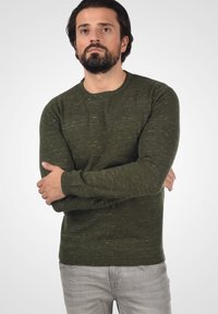 Khaki groene sweater van textuurgebreide stof, met een ronde hals en ribgebreide manchetten. Draagt met lichtgrijze jeans.