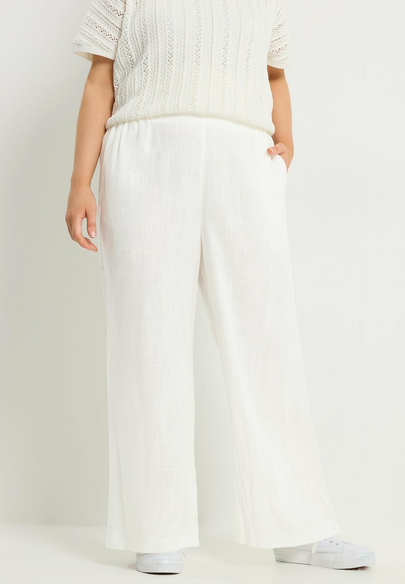 Osoba ubrana w luźne białe lniane spodnie, biały krótki sweter z dzianiny oraz białe sneakersy, stojąca z jedną ręką w kieszeni.