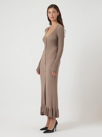 Vrouw die staat in een lange taupe geribde gebreide jurk met knopen aan de voorkant en zwarte high heels op een witte achtergrond.