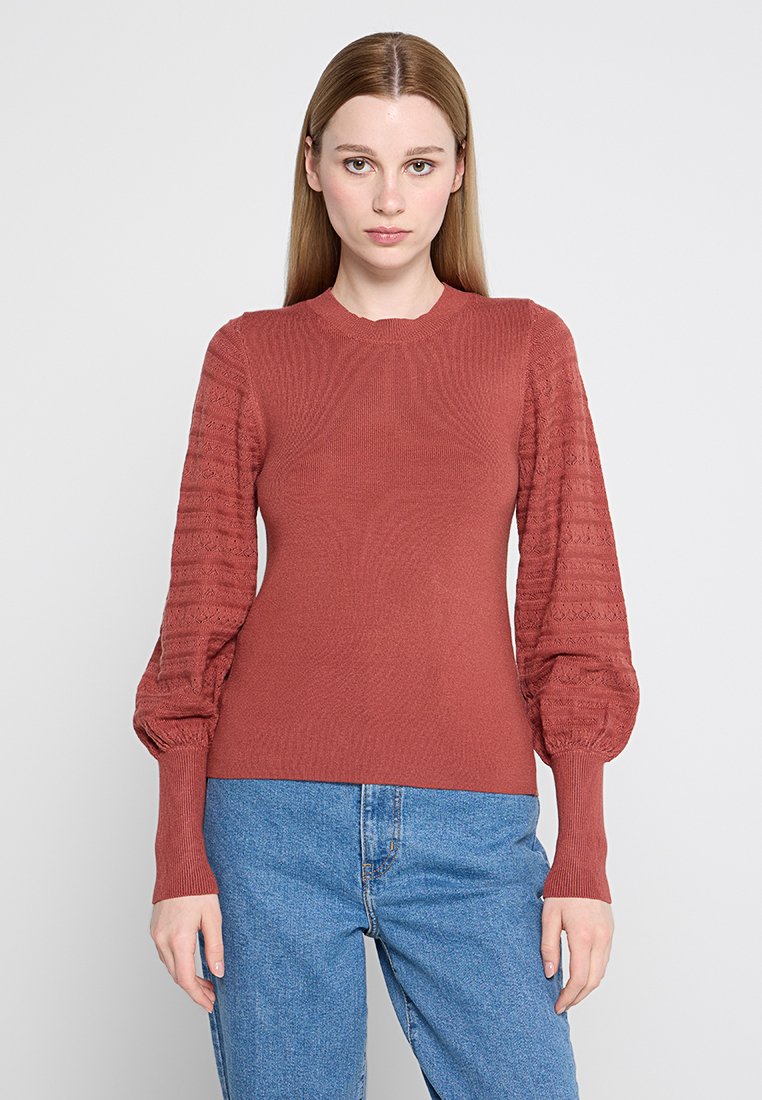 Pull en Maille couleur rouille avec un corps côtelé et des manches bouffantes texturées. Doté d'un col rond et de poignets ajustés. Associé à un jean bleu.