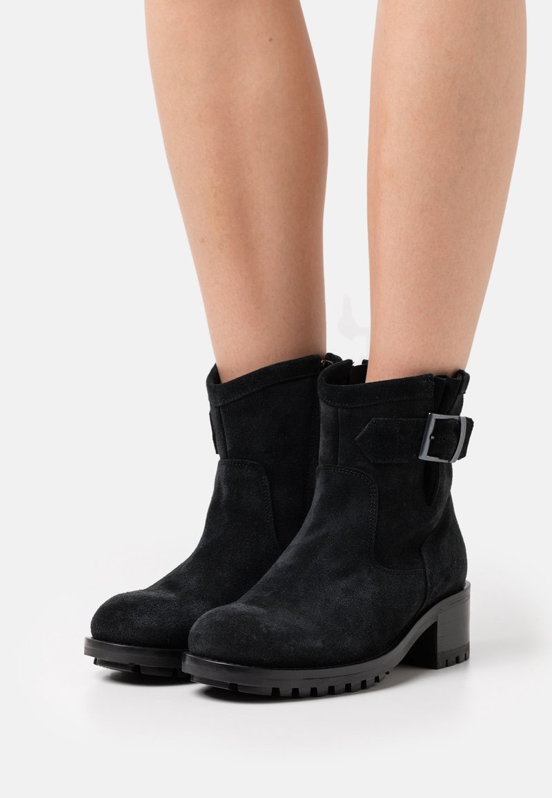 Botines de ante negro con tacón grueso, presentan un detalle de hebilla y una textura suave. El diseño incluye un cuello plisado para mayor detalle.