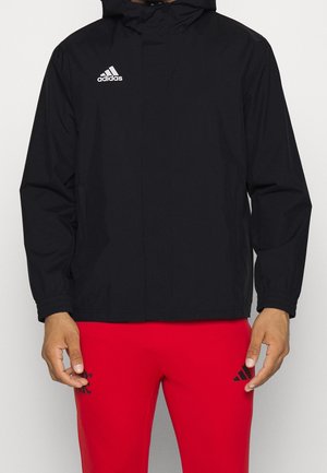 Homme portant une veste noire Adidas avec capuche et un pantalon Adidas rouge sur un fond clair uni.