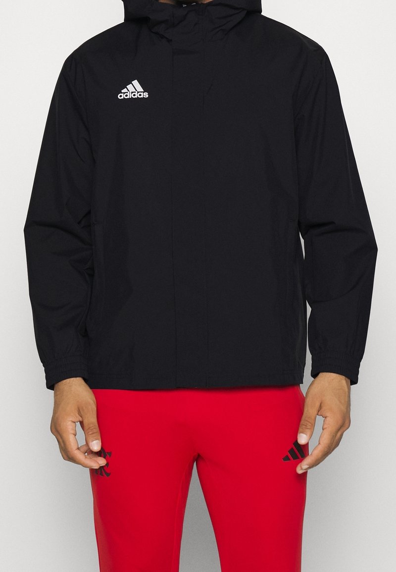 Homme portant une veste noire Adidas avec capuche et un pantalon Adidas rouge sur un fond clair uni.