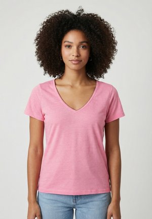 Donna con capelli ricci che indossa una maglietta rosa con scollo a V e maniche corte e jeans azzurri, in piedi davanti a uno sfondo chiaro e semplice.