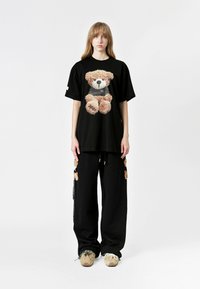 Camiseta negra de gran tamaño con un gráfico de un oso de peluche, combinada con pantalones negros que tienen acentos de oso de peluche. Ajuste casual con telas texturizadas.