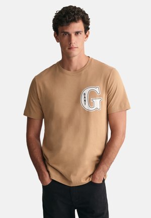 T-shirt en coton marron avec une grande lettre blanche "G" et le mot "GANT" en noir, encolure ronde, manches courtes et coupe décontractée.