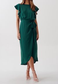 Abito avvolgente verde in tessuto leggero, con maniche corte a flutti, cintura da annodare e uno spacco laterale. Completato con sandali con cinturini.