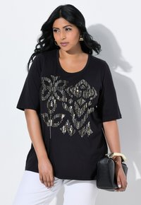 Una mujer con cabello largo y oscuro, vestida con una camiseta negra con un diseño metálico, sostiene un bolso negro mientras está de pie frente a un fondo claro.