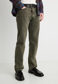 Άνδρας που φοράει χακί παντελόνι τζιν, μαύρη ζώνη, μαύρα δερμάτινα μοκασίνια και καρό πουκάμισο, με το ένα χέρι στην τσέπη, στέκεται σε λευκό δάπεδο.